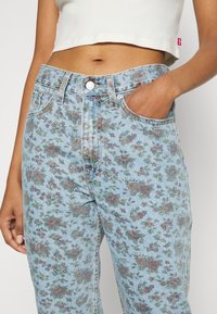 Jeans de color azul claro con estampado floral que presentan una cintura alta, cinco bolsillos y herrajes metálicos, con una ligera textura desgastada.