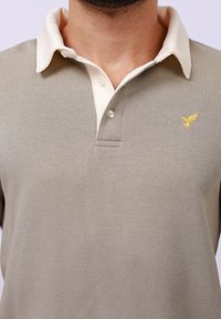 Beige polo shirt with a cream-colored collar, featuring a three-button placket and a small yellow embroidered bird logo on the chest.