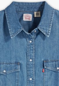 Helle blaue Denim-Bluse mit einem spitzen Kragen, Druckknöpfen, zwei Brusttaschen und einem kleinen roten Etikett-Akzent an der Seite.