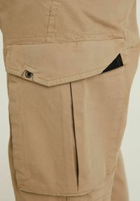 Beżowe spodnie cargo z dużą kieszenią boczną z klapą i przeszyciami. Materiał wydaje się trwały i ma gładką fakturę.