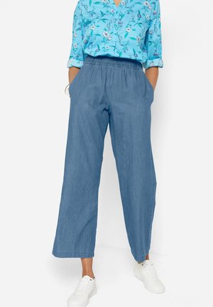 Femme portant un pantalon en jean bleu ample avec une taille élastique et des baskets blanches, associé à une chemise bleue fleurie boutonnée aux manches retroussées.