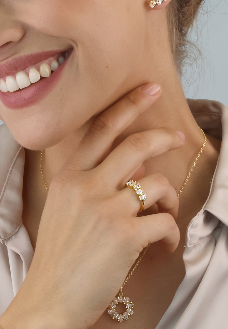 Gold jewelry set featuring a ring with five clear gems, a circular pendant, and matching earrings. Smooth texture, elegant design.