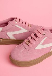 Roze suede tenisice s bijelim detaljima, okruglim vezicama i metalnim elementima na jeziku. Imaju nisku gumenu podlogu i teksturiranu površinu.