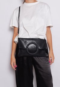 Zwarte leren handtas met een ronde accenten, slank ontwerp en een afneembare schouderband, gedragen met een wit T-shirt en donkere broeken.