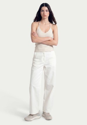 Young woman with long dark hair standing with arms crossed, wearing a beige striped halter top, white wide-leg pants, and beige sneakers.