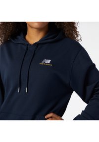Felpa blu navy con cappuccio regolabile, tasca a marsupio e un piccolo logo "New Balance" ricamato in argento e oro. Tessuto morbido.