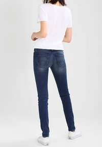 Jeans de denim azul con un corte ajustado, que presentan bolsillos traseros y detalles de desvanecimiento sutil, combinados con una camiseta blanca y zapatillas blancas.