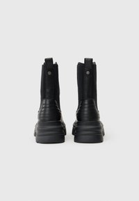 Botas de tornozelo pretas com um cabedal em couro texturizado e tecido elástico, apresentando uma sola grossa e costura proeminente no calcanhar.