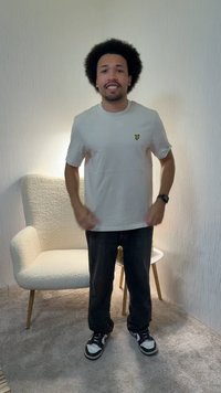 Νεαρός με κούρεμα αφρο, φορώντας λευκό μπλουζάκι, μαύρο παντελόνι και αθλητικά παπούτσια, στέκεται χαμογελώντας σε ένα δωμάτιο με χαλί, με μια λευκή πολυθρόνα και ένα μικρό τραπέζι.