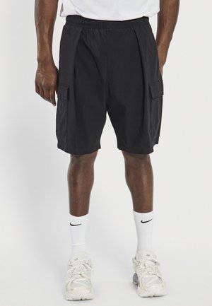 Hombre vistiendo pantalones cortos cargo negros, calcetas blancas de Nike y zapatillas blancas, de pie frente a un fondo blanco liso.