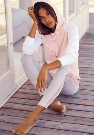 Helle rosa und weiße Kapuzenjacke mit Kordelzügen, kombiniert mit grauen Leggings, die mit einem pinken Logo-Streifen verziert sind. Hintergrund aus Holzfußboden.