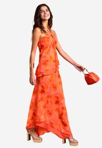 Maxikleid in leuchtendem Orange mit floralen Mustern, geschichtetem Design, dünnen Trägern und fließender Silhouette. Kombiniert mit Plateausandalen und einer Handtasche.