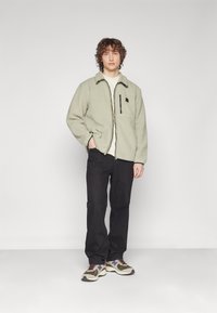Giacca in pile con zip di colore verde chiaro, dotata di una tasca sul petto nera e una zip a contrasto. Indossata sopra una camicia chiara e abbinata a pantaloni scuri e sneakers.