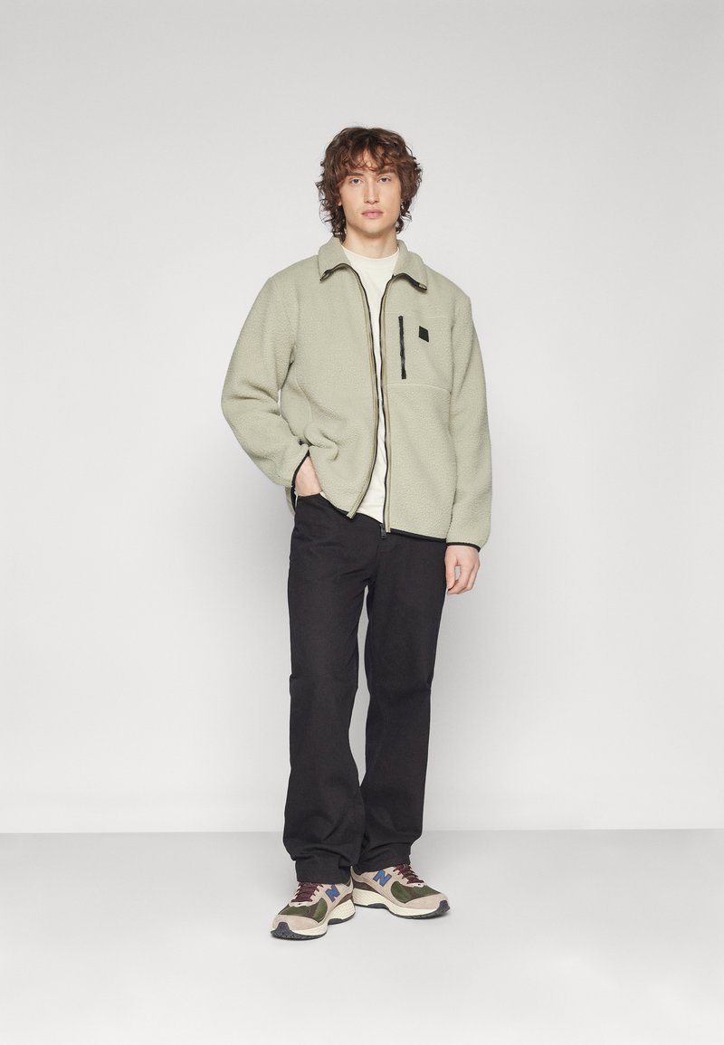 Fleecepiké jacka i ljusgrön, med en svart bröstficka och kontrasterande dragkedja. Bärs över en ljus skjorta och matchas med mörka byxor och sneakers.