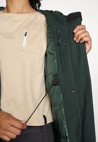 Veste vert foncé avec une poche intérieure zippée, portée par-dessus une chemise texturée beige avec une poche poitrine. Cordon de serrage ajustable à l'intérieur.