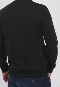 Uomo che indossa una felpa nera semplice a maniche lunghe e jeans blu scuro, visto di spalle su uno sfondo neutro.