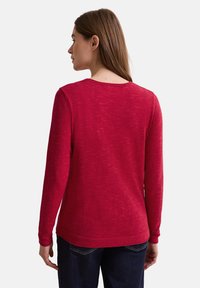 Roter Pullover mit langen Ärmeln, strukturiertem Finish, Rundhalsausschnitt und gerippten Bündchen, kombiniert mit dunkelblauen Jeans für einen Kontrast.