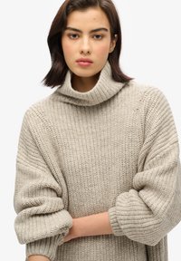 Suéter de punto beige claro con cuello alto, detalles acanalados, mangas de gran tamaño y un patrón texturizado en todo su diseño.