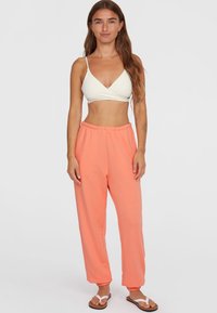 Lichtgewicht, witte, driehoekige bralette gecombineerd met felgekleurde koraalkleurige sweatpants met elastische tailleband. Glad van textuur, taps toelopende boorden bij de enkels, gedragen met sandalen.