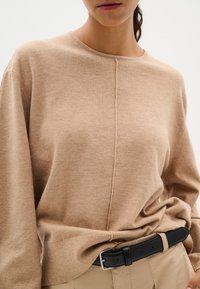 Maglione beige realizzato in tessuto morbido, caratterizzato da un collo rotondo, maniche lunghe e una cucitura centrale; abbinato a una cintura in pelle nera.