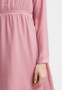 Lyserød langærmet kjole lavet af glat stof, med elastisk talje og manchetærmer. Enkel design med en feminin silhuet.