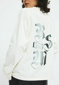 White long-sleeve sweatshirt with a relaxed fit, featuring a large black and gray graphic design on the back, including stylized letters and numbers.