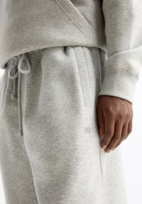 Nærbillede af en person iført lysegrå sweatpants og en matchende sweatshirt, med en hånd hvilende ved lommen på bukserne.