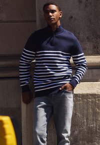 Suéter de lana navy con rayas horizontales blancas, cuello con media cremallera y puños acanalados. Combinado con jeans de color azul claro. Fondo urbano.