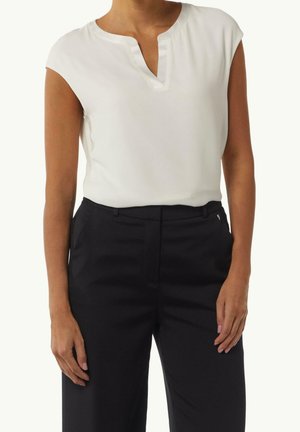 Femme portant un haut blanc sans manches avec un décolleté en V et un pantalon noir taille haute, debout les bras détendus le long du corps.