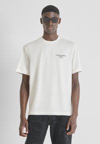 Camiseta de algodón blanca de manga corta y cuello redondo, con el texto "ANTHONY MORATO" en negro en el pecho, combinada con jeans oscuros y gafas de sol.