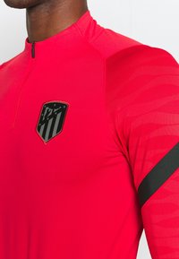 Camiseta atlética roja con media cremallera, mangas largas y una banda de acento negra. Presenta un logo negro con rayas y estrellas en el pecho.