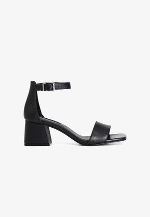Sandale noire à talon avec une bride de cheville et une boucle en argent. Elle possède un talon carré et une tige en cuir lisse au design minimaliste.
