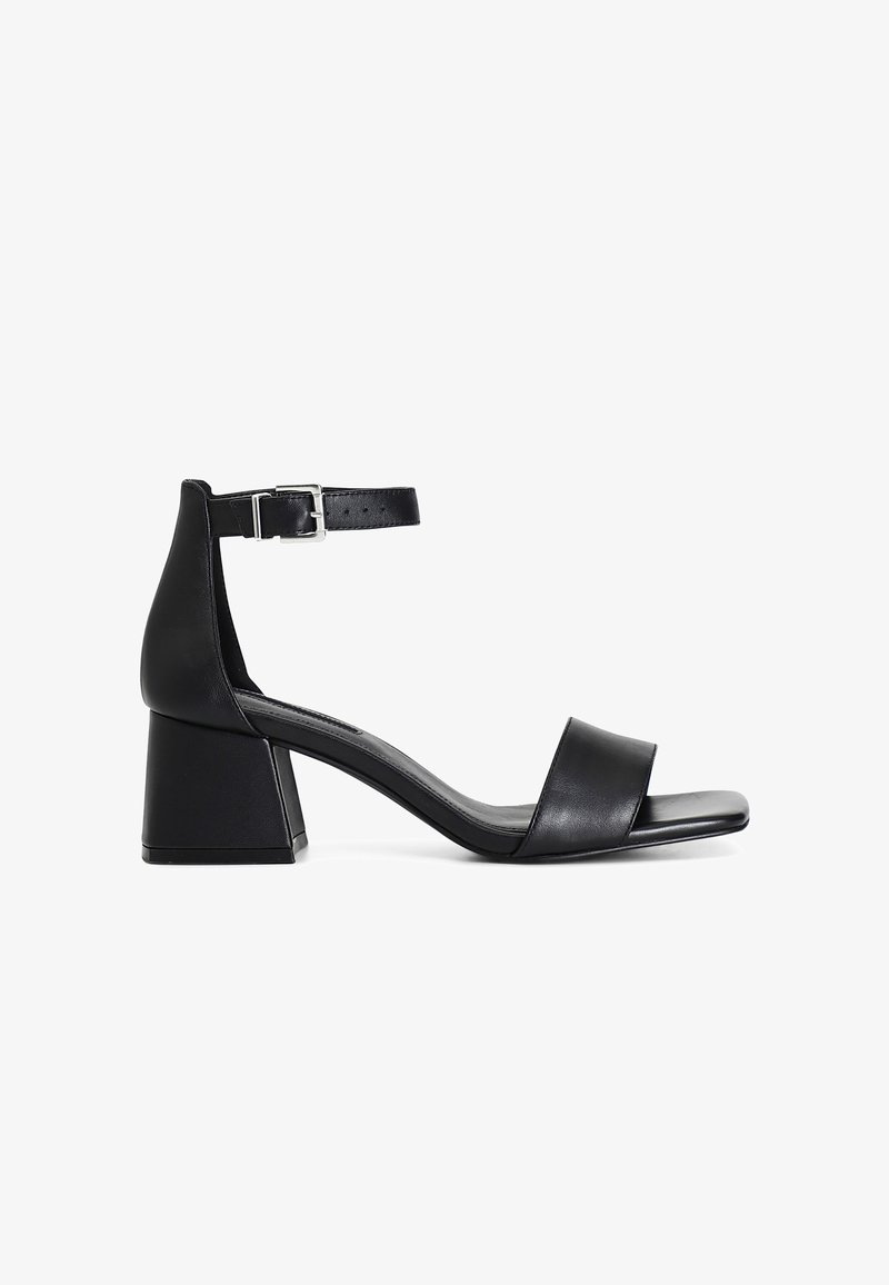 Musta korkokenkä nilkkaremmin ja hopean soljen kanssa. Varustettu tukeva korko ja sileä nahkainen päällinen, jossa on minimalistinen muotoilu.