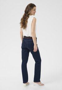 Jean en denim de couleur bleu foncé avec une coupe droite, dotés de poches arrière et de coutures contrastées, associés à un haut sans manches clair.