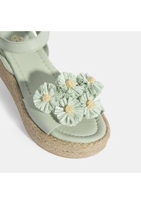 Hellgrüner Leder-Sandalette mit floralen Verzierungen, geflochtenem Jute-Plateau und weicher, glatter Textur. Verstellbarer Knöchelriemen enthalten.