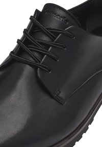 Chaussure en cuir noir avec une finition lisse, présentant un bout rond, des lacets plats, une languette et une semelle texturée. Marquage subtil sur le côté.