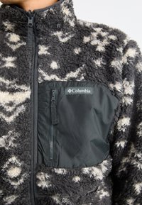 Fleece-Jacke mit einem grauen und cremfarbenen Muster, einer Reißverschlusstasche mit einem schwarzen Stoffpanel und einem hohen Kragen. Markenlabel sichtbar.