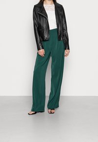 Veste de motard en cuir noir, haut en dentelle blanche et pantalon large vert. La tenue présente des textures lisses et une silhouette fluide.