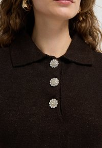 Žena v tmavém pleteném svetr s límečkem a třemi dekorativními kamínkovými knoflíky vpředu.