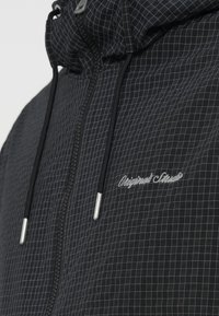 Μαύρο μπουφάν με φερμουάρ και σχέδιο σε μορφή πλέγματος, με κουκούλα με κορδόνι, ασημένια εξαρτήματα και κεντημένο λογότυπο στο στήθος.