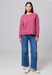 Sweatshirt rose avec col rond et détails aux épaules, associé à un jean bleu à jambe large. Des baskets blanches ajoutent une touche décontractée.