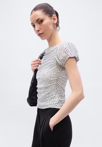 Λευκό μπλουζάκι με πουά, κοντά φουσκωτά μανίκια, ραμμένη υφή και εφαρμοστή σιλουέτα, σε συνδυασμό με μαύρο παντελόνι ραμμένο κατά παραγγελία.