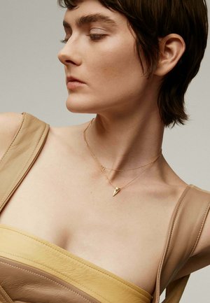 Collares de oro en capas con un pequeño colgante triangular, combinados con un top de cuero beige que presenta un escote asimétrico.