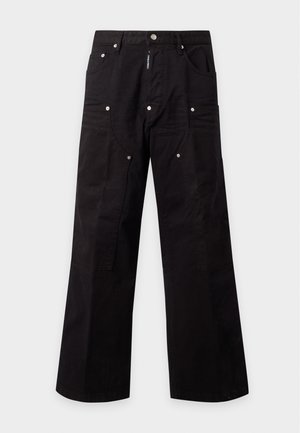 Jeune homme portant un haut blanc sans manches, un pantalon noir à jambes larges avec ceinture et des chaussures noires, debout devant un fond blanc uni.