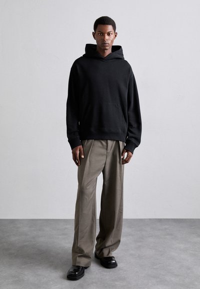 Haut en coton noir avec poche kangourou à l'avant, associé à un pantalon beige à coupe large et des bottes noires brillantes. Tissu texturé et silhouette décontractée.