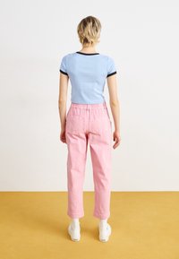 Persona con pelo corto rubio que lleva una camiseta corta de color azul claro, pantalones de cintura alta rosa, calcetines blancos y zapatillas blancas, de pie sobre un suelo amarillo.