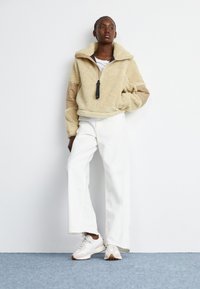 Pull en polaire beige avec une demi-fermeture éclair, manches contrastantes couleur tan et col haut ; associé à un pantalon large blanc et des baskets beiges.