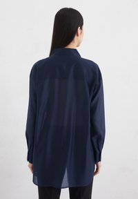 Marineblauwe, lichte shirt met een kraag en knopenmanchetten. Heeft een semi-doorzichtige achterpanneel en lange mouwen. Op maat gemaakte, relaxte pasvorm.
