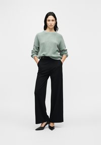 Jasnozielony sweter dzianinowy o luźnym kroju, zestawiony z czarnymi spodniami o szerokich nogawkach z ozdobnym guzikiem. Całość dopełniają czarne szpilki na spiczastym nosku.