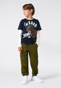 Chłopiec stoi, patrząc w lewo, ubrany w wielowarstwowy granatowy T-shirt z grafiką dinozaura, białe długie rękawy, oliwkowe spodnie cargo i białe trampki.